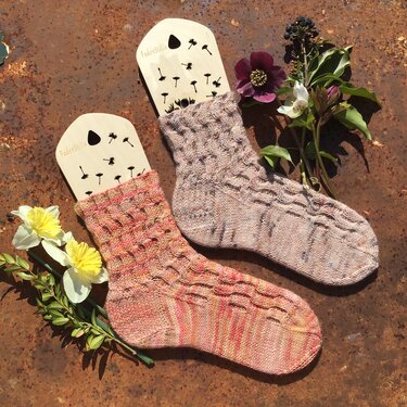 Zwei gestrickte Damensocken auf Sockenbrettchen, eine in Rosé-Mischfarben und eine in Beige-Mischfarben, beide mit strukturierter Wellenoptik, auf rostigem Untergrund mit Blumen.