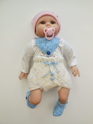 Babypuppe in gestricktem Baby‑Set: dreieckiges Lätzchen, kurze Hose mit Kordelzug und Zierknöpfen sowie passende Söckchen in Pastellfarben.