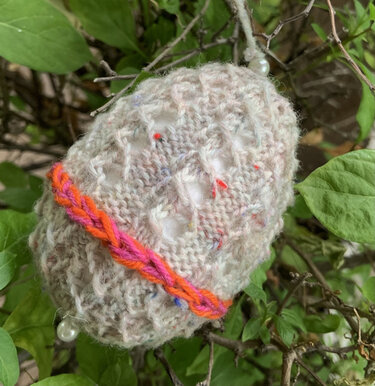Gestricktes Osterei aus hellem Fleckgarn mit orange-pinkem Zierband und Perlen, hängend an Zweigen