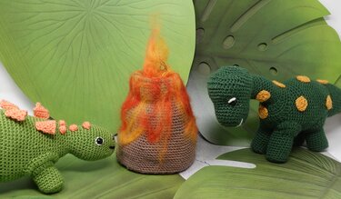 Zwei gehäkelte grüne Amigurumi-Dinosaurier und ein gehäkelter Vulkan mit orange-roter Filzflamme vor großen grünen Blättern