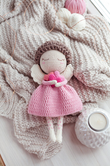 Crocheted angel doll in a pink dress holding small pink hearts, on a beige knitted blanket with yarn and fairy lights.