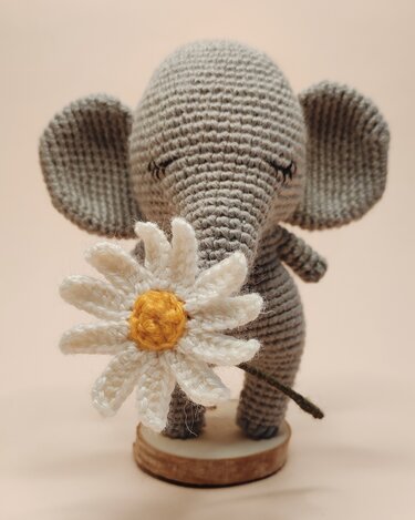 Grey crocheted amigurumi elephant holding a white daisy on a small wooden base.