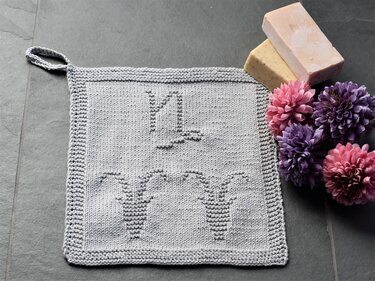 Light grey square knitted washcloth with a raised watering can and three potted plants motif, with a hanging loop, photographed beside soap bars and pink and purple flowers on a dark surface.