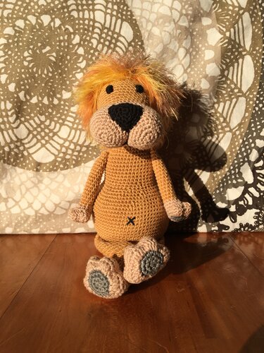 Gehäkelter Amigurumi-Löwe mit orange-gelber Mähne und schwarzer Nase, sitzend auf Holzfußboden vor gemustertem Stoff.