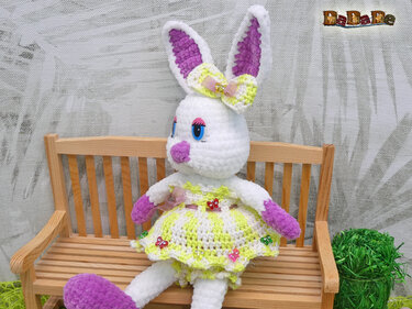 Sitzender Amigurumi-Hase mit lila Pfoten, Schleife und gelb-weißem Kleid auf Holzbank