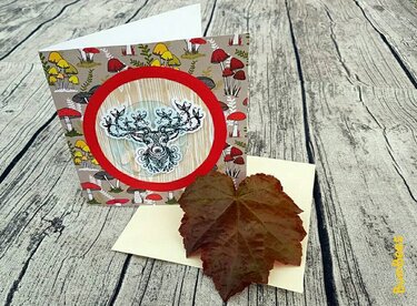 Greeting card with ornamental deer head in a red circle and mushroom patterned background, shown on a wooden surface with an envelope and dried leaf.