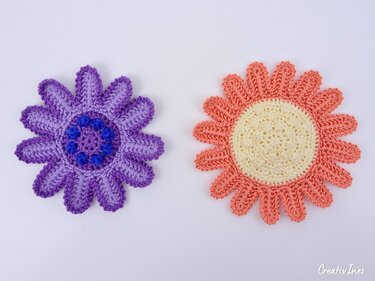 Nahaufnahme zweier gehäkelter, blumenförmiger Untersetzer in Violett und Koralle, mit sichtbaren Maschenreihen