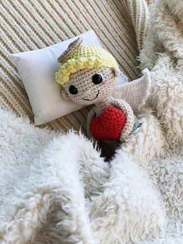 crocheted amigurumi angel doll with yellow hair, halo, white wings holding a red heart on a pillow and fluffy blanket