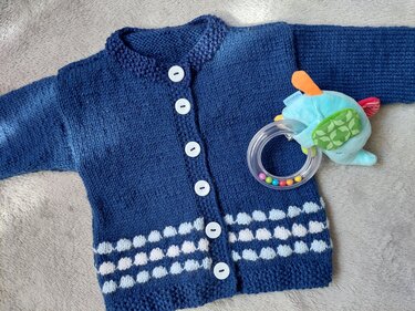 Dunkelblaue gestrickte Babyjacke mit weißen Knöpfen und weißem Noppenmuster, daneben ein weiches Spielzeug.