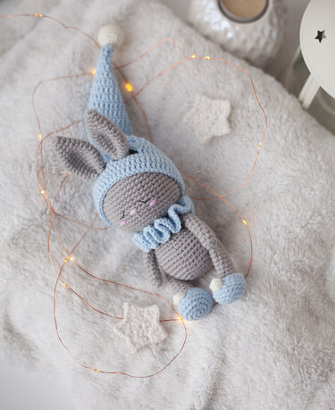 Grey crocheted amigurumi bunny wearing a light blue sleep hat, ruffled collar and booties, lying on a soft white blanket with fairy lights and small star decorations.