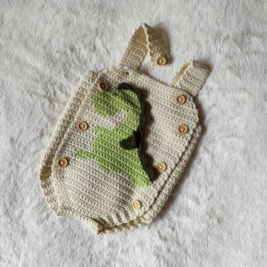 Cream crocheted baby romper with green dinosaur appliqué and wooden buttons on a soft white surface.