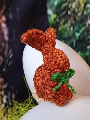 Handgehäkelter brauner Hase mit grüner Schleife auf weißem Ei vor unscharfem, grünem Hintergrund