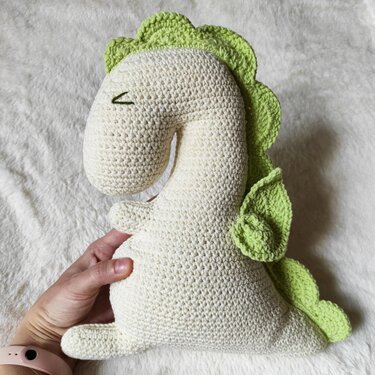 Off-white crocheted dinosaur plush with green spikes and wings, held by a hand on a white fluffy background.
