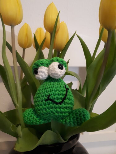 Gehäkelter grüner Frosch mit großen weißen Augen und einem Lächeln zwischen gelben Tulpen und grünen Blättern.