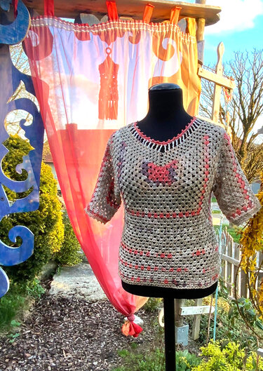 Gehäkeltes Damen‑Shirt in Rosa und Grau mit V‑Ausschnitt und Schmetterlingsmotiv auf Mannequin vor Gartenkulisse