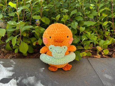 Orangefarbenes gehäkeltes Küken mit mintgrünem Frosch-Schwimmring auf Gehweg vor Pflanzen