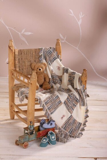 Holzstuhl mit karierter Babydecke, Teddybär, Spielzeug-Lkw und zwei Paar Babyschuhe auf hellem Boden.