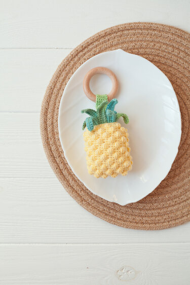 Crocheted yellow pineapple baby teether with green leaves on a wooden ring, placed on a white plate over a woven mat