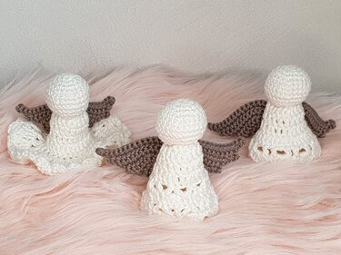 Drei weiße, gehäkelte Amigurumi-Engel mit taupefarbenen Flügeln auf rosa Plüsch