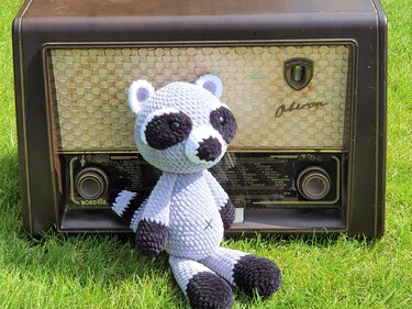 Gehäkelter Amigurumi-Waschbär in Grau und Schwarz sitzt auf Gras vor einem alten Radio.