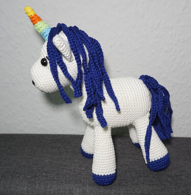 Gehäkeltes weißes Einhorn mit blauer Mähne, regenbogenfarbenem Horn und schwarzen Augen, seitliche Ansicht