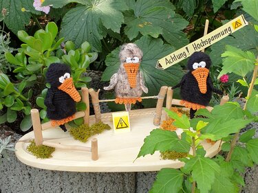 Drei freche Amigurumi-Vögel mit orange Schnäbeln auf Holzaufsteller und Warnschild im Garten