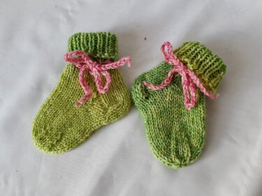 Zwei grüne, gestrickte Babysocken mit geripptem Bündchen und rosa Bindeband auf hellem Untergrund
