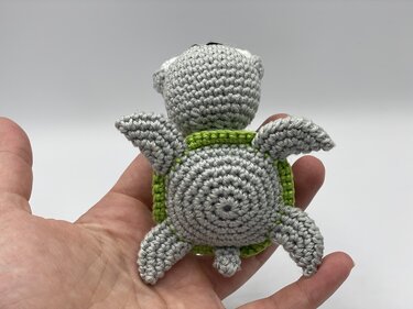 Hand holding a small crocheted turtle with a grey body and green shell, viewed from the back.