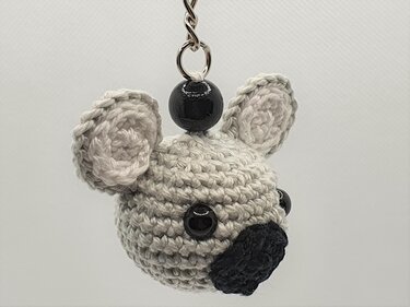 Nahaufnahme eines gehäkelten hellgrauen Koala‑Kopf‑Schlüsselanhängers mit schwarzer Holzperle, glänzenden schwarzen Augen und schwarzer Nase an Metallring.