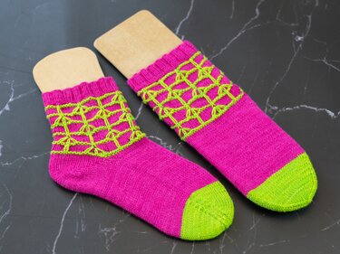 Zwei gestrickte Socken in Pink mit neongrünen Zehen und gitterartigem Lochmuster auf dunklem marmoriertem Untergrund.