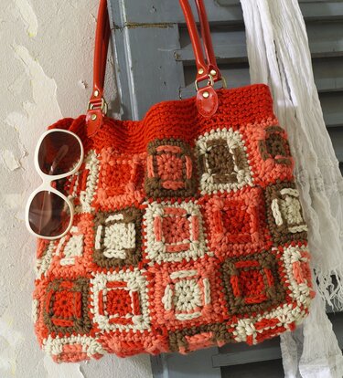 Große gehäkelte Tasche mit Granny-Square-Mustern in Orange, Braun und Creme, an der eine weiße Sonnenbrille hängt