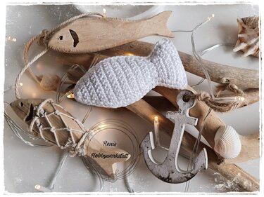 Nahaufnahme eines weißen gehäkelten Amigurumi-Fischs mit sichtbarer Maschenstruktur, arrangiert mit Muscheln und Holzanker