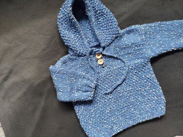 Blauer gestrickter Kapuzenpullover für Babys mit Kapuze und zwei Holzknöpfen auf dunklem Hintergrund.