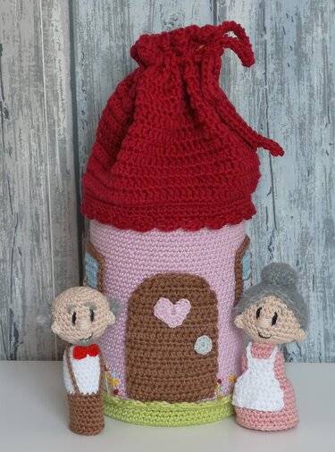 Detailaufnahme eines gehäkelten Hauses mit rosafarbenen Wänden und rotem Beuteldach mit Zugband, daneben zwei Amigurumi-Handpuppen (Großmutter und Großvater).