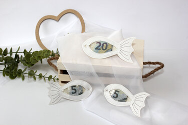 Weiße Fisch‑Anhänger mit Sichtfenster und Geldscheinen auf Holzkiste, Herzdekoration und Grünzweigen