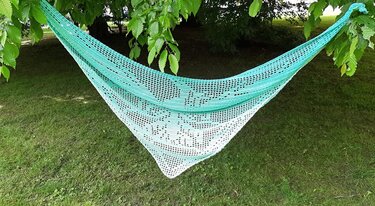 Gehäkeltes Dreieckstuch in Mintgrün und Weiß mit feinem Spitzenmuster, zwischen Baumzweigen aufgehängt über einer Wiese.