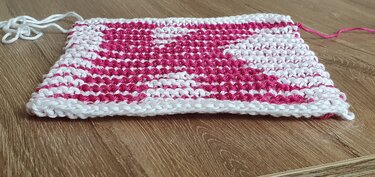 Gestrickte Maschenprobe in Weiß und Pink mit sichtbarer Seitenkante auf Holzboden
