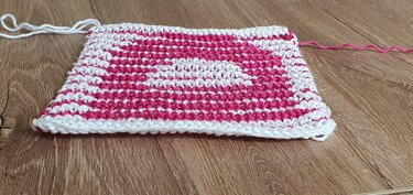 Rechteckige Maschenprobe mit weißen und pinken Streifen auf Holzuntergrund