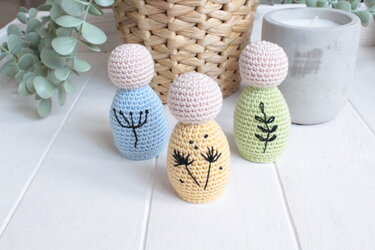 Drei kleine pastellfarbene Amigurumi-Püppchen (Blau, Gelb, Grün) mit schwarzen Stickmotiven