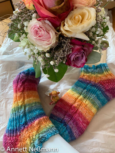 Paar bunter gestrickter Socken mit Regenbogenstreifen neben Rosenstrauß und kleiner Dekofigur