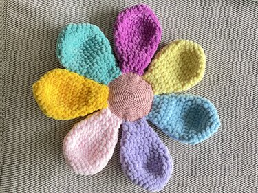 Crocheted flower-shaped baby snuggle blanket with seven colorful plush petals and a round pink center on a textured gray background