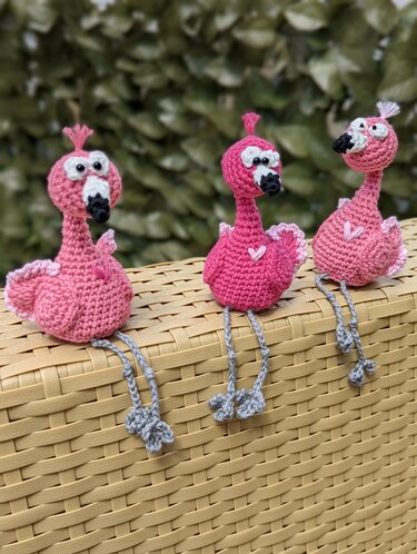 Drei gehäkelte rosa Flamingos mit grauen Beinen auf geflochtenem Korb
