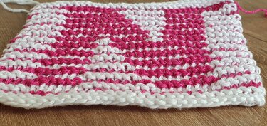 Gestrickte Musterprobe in Pink und Weiß mit wiederholtem Herz- und Rautenmuster auf Holzuntergrund.
