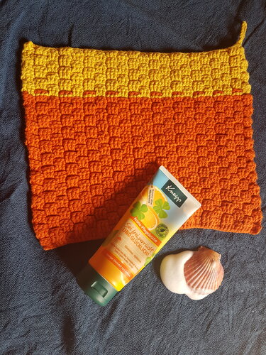 Rechteckiger, handgehäkelter Waschlappen in Gelb und Orange mit erhabenem Reliefmuster auf dunklem Tuch, daneben eine Kosmetiktube und eine Muschel.
