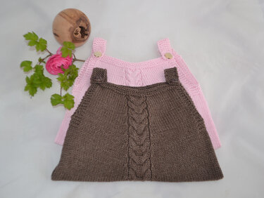 Zwei gestrickte Baby-Latzhosen in Rosa und Braun mit Zopfmuster und Knopfträgern, dekoriert mit Blume und Holzobjekt.