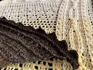 Close-up of a dark brown crocheted triangle shawl with scalloped edge layered on a cream open-mesh crochet fabric