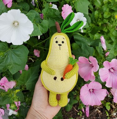 Hand holding a yellow pear-shaped amigurumi with a bunny-like face and green top leaves, hugging a small crocheted carrot in a flower garden.
