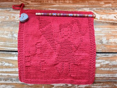 Fuchsiafarbenes, quadratisches Stricktuch mit eingestricktem Seepferdchen-Herz auf rustikaler Holzoberfläche; Bleistift, herzförmiger Knopf und Aufhängeschlaufe sichtbar.