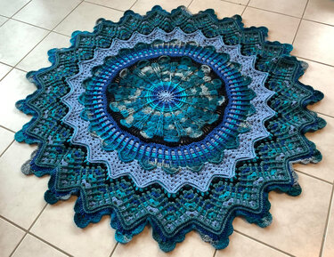 Runde gehäkelte Decke in Blau- und Türkistönen mit komplexem Mandala-Muster und gewelltem Rand auf hellem Boden.