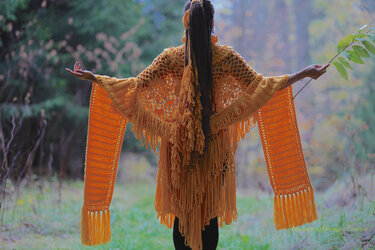 Rückenansicht einer Person mit senfgelbem, gestricktem Schal mit langen Fransen und offenem Muster im Wald